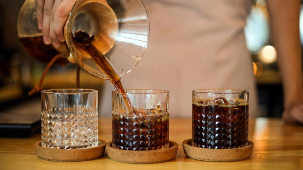 A man brewing Chemex coffee at home. He's pouring 3 glasses with Chemex coffee for his guests