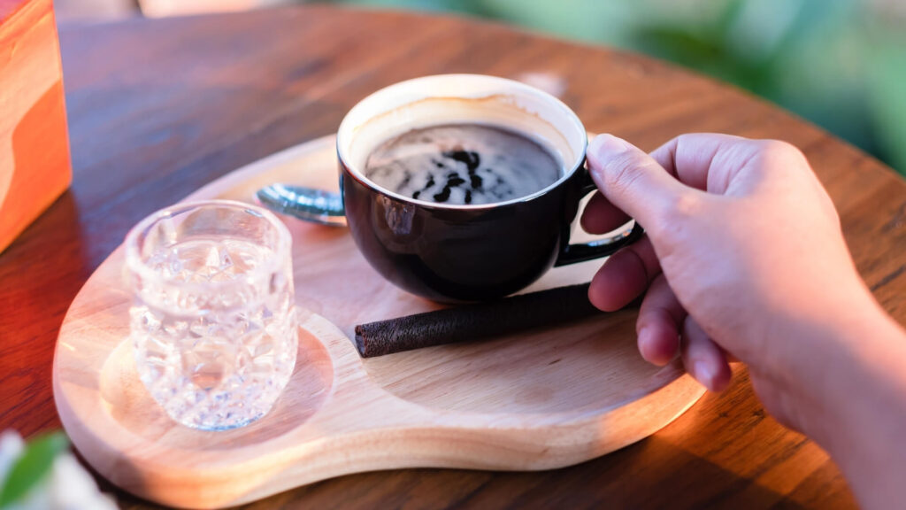 Woman's hand holding a cup that is full of red eye coffee. There's a glass of water visible too, water is used o clean coffee buds before tasting the coffee