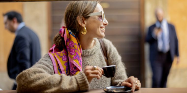 Italian woman drinking espresso in a cafe. She is wearing glasses, colorful scarf and a light-brown pull over