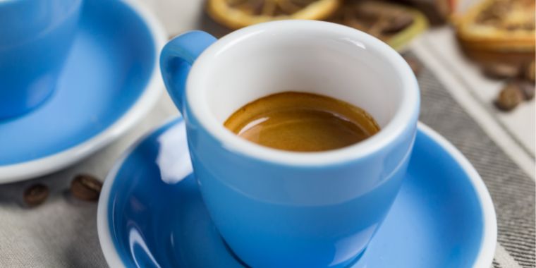 A blue espresso cup filled with a shot of rich, dark espresso. The cup is sitting on a saucer. The background is blurred, with hints of brown and gray colors visible