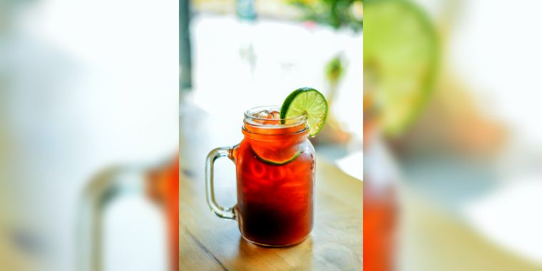 A tall clear jug filled with ice cubes and sparkling water, topped with a shot of espresso and a slice of lime. The drink has a rich brown color with bubbles rising to the top.